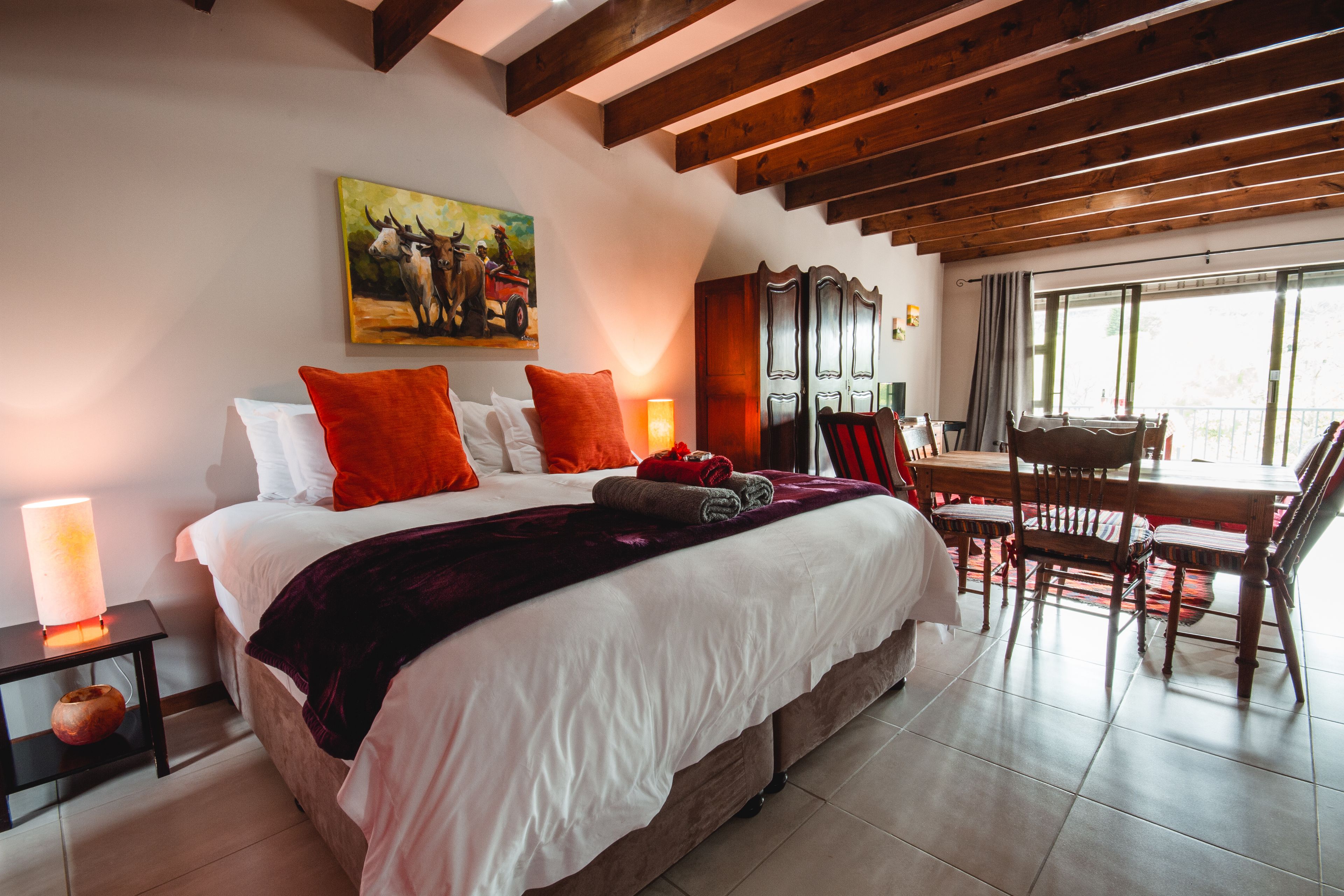 A bedroom with a large bed, a painting on the wall, and a dining area with chairs and a table.
