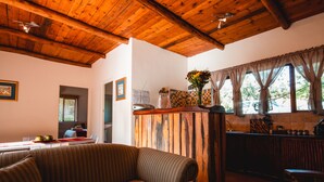A cozy room with a wooden ceiling, a sofa, a dining table with apples, and a kitchen area with a wooden counter and window with curtains. - Wildebraam Berry Estate (Swellendam)