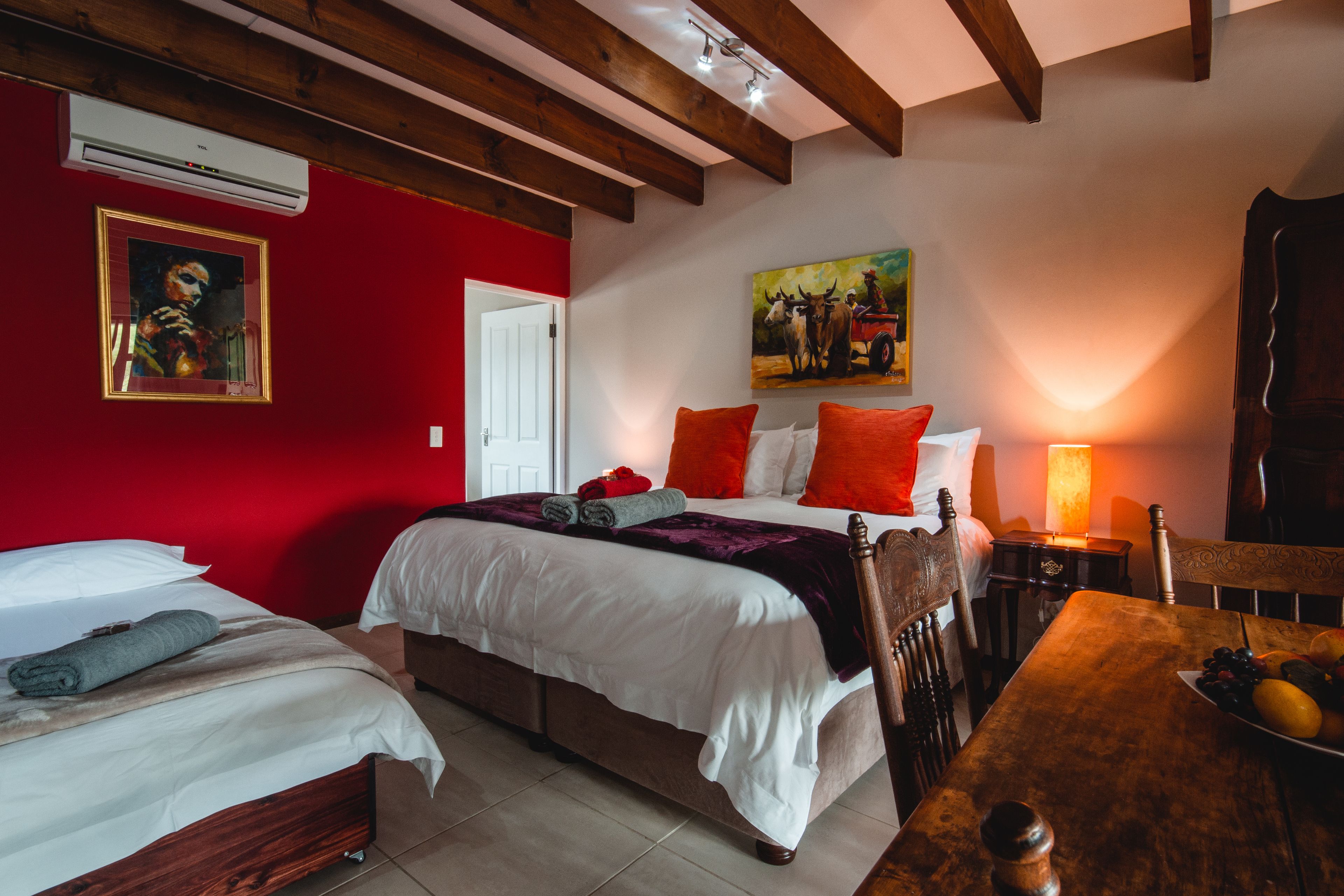 A hotel room with two beds, a wooden table with fruit, a painting, and a red wall.