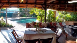 An outdoor dining area with a pool, wooden furniture, and tropical plants. - Polokwane Place (Polokwane)