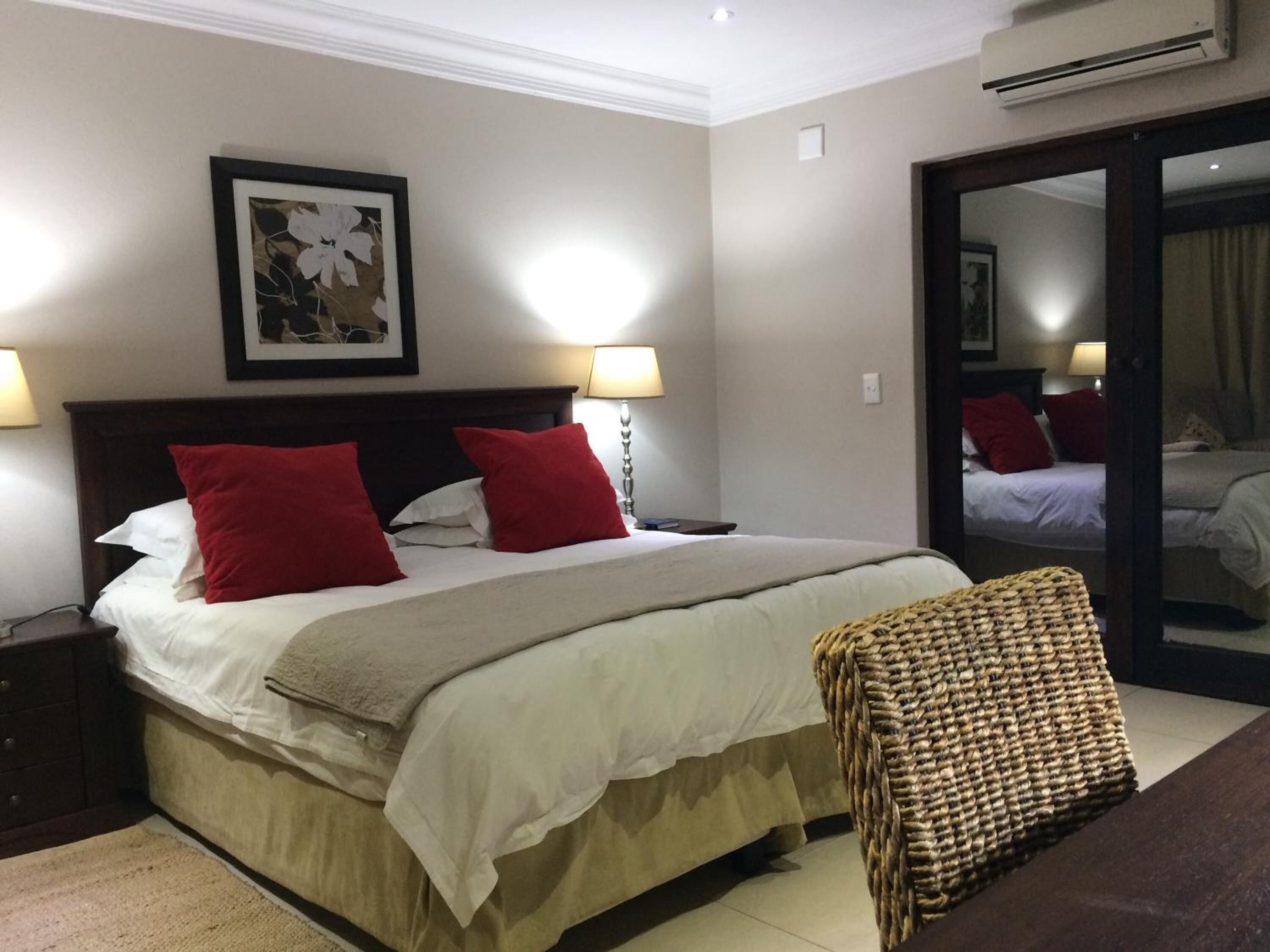 A neatly made bed with red and white pillows, a bedside lamp, and a woven bench.