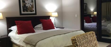 A neatly made bed with red and white pillows, a bedside lamp, and a woven bench.