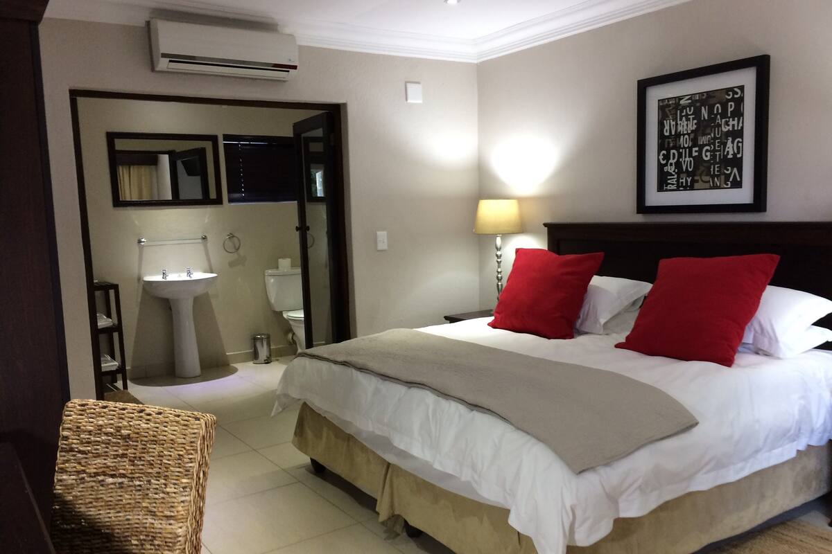 A hotel room with a bed, a bedside table, a lamp, a framed picture on the wall, and an air conditioning unit.