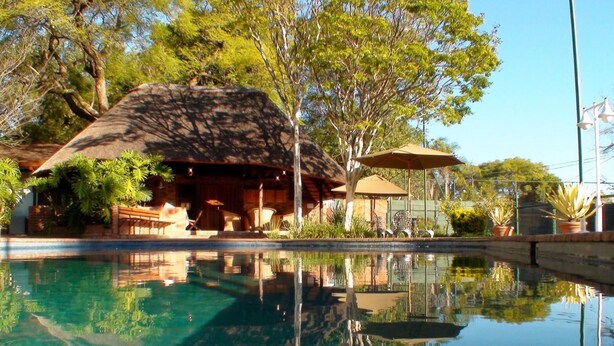 A clear pool with a reflection of a thatched-roof structure and surrounding trees. - Polokwane Place (Polokwane)