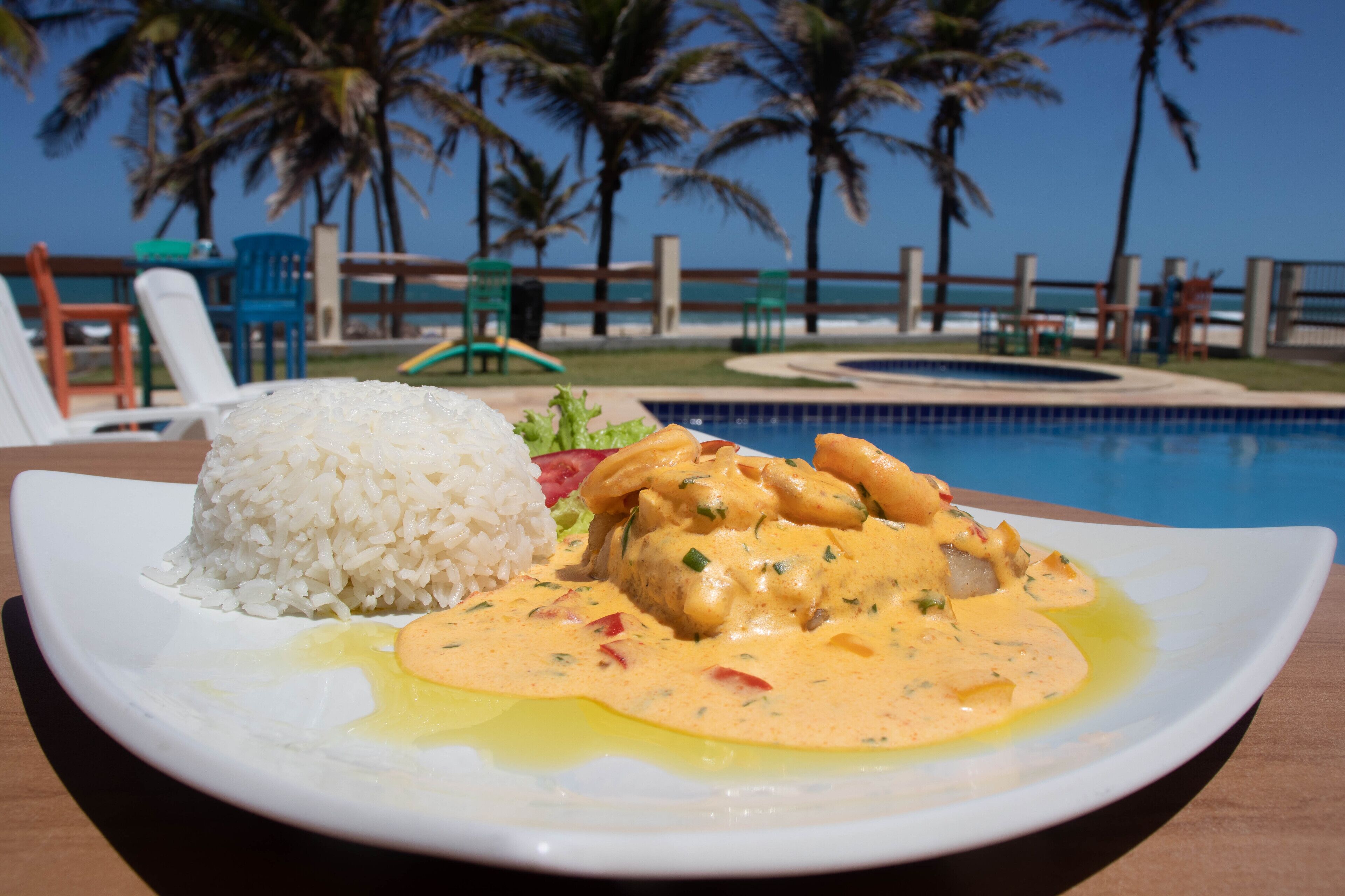A plate with white rice and a creamy dish, possibly curry, on a wooden table by a poolside with palm trees and lounge chairs in the background.