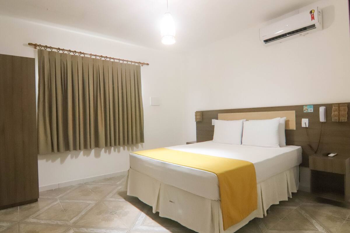 A hotel room with a bed, curtains, an air conditioning unit, a bedside table, and a nightstand.
