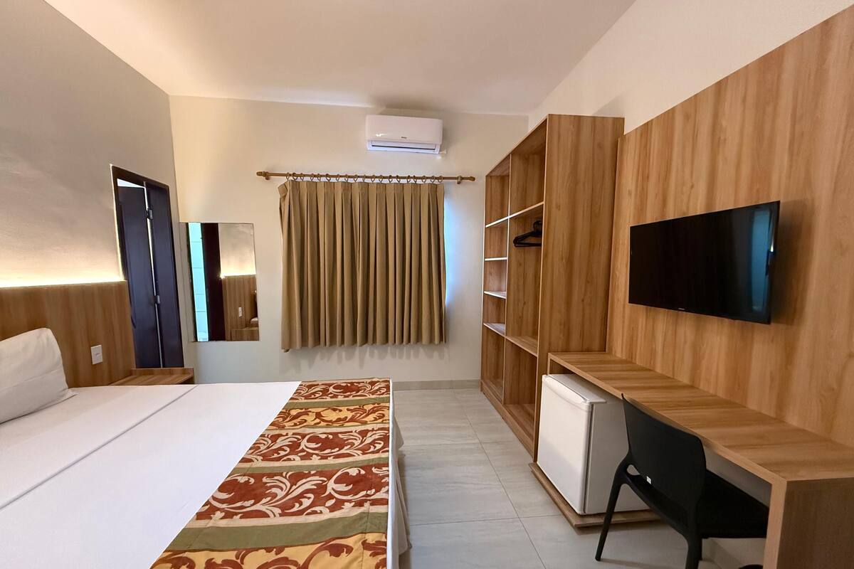 A hotel room with a bed, a desk, a TV, a wardrobe, and an air conditioning unit.