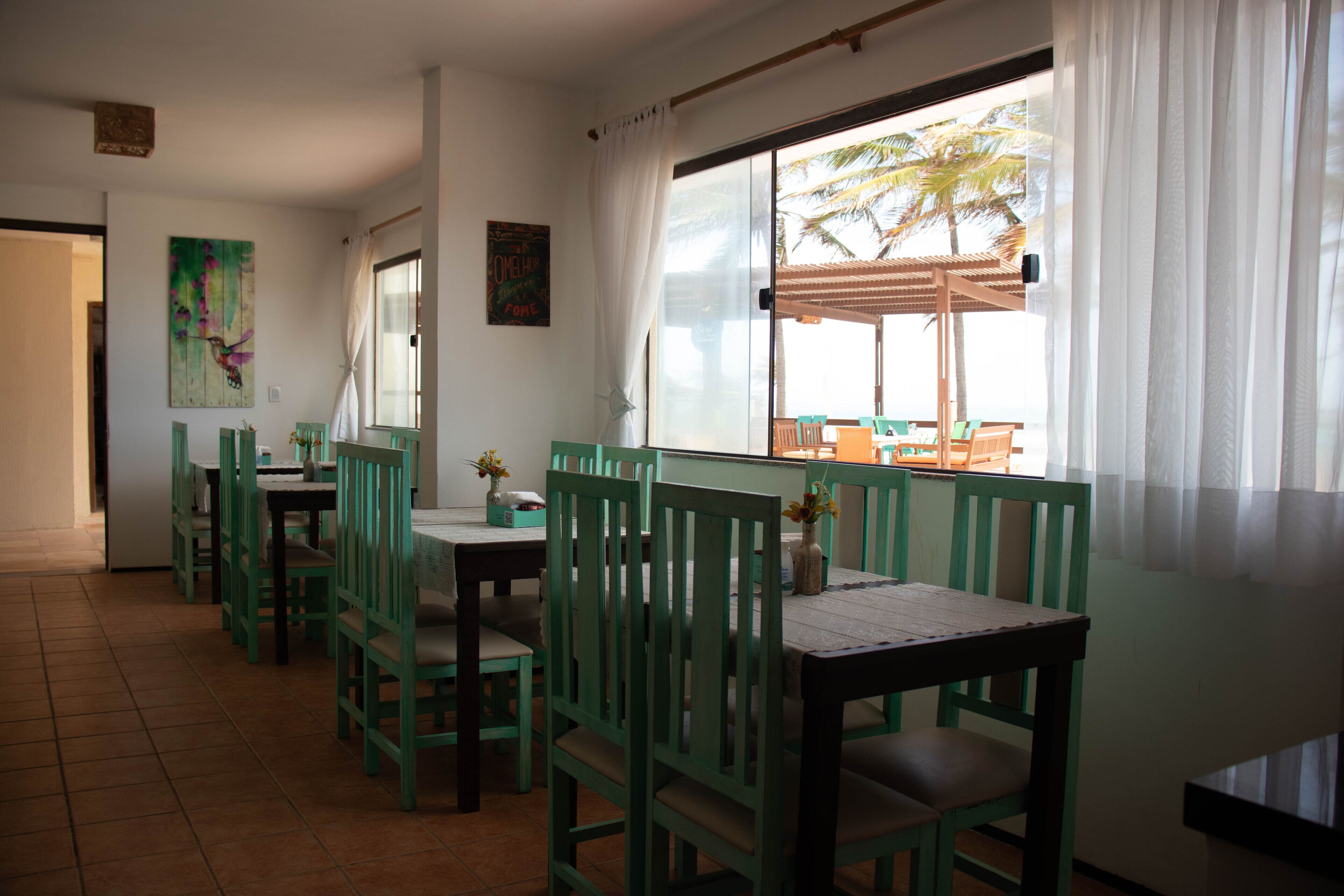 Un comedor con sillas y mesas verdes, un ventanal con vistas a palmeras y un cuadro en la pared.