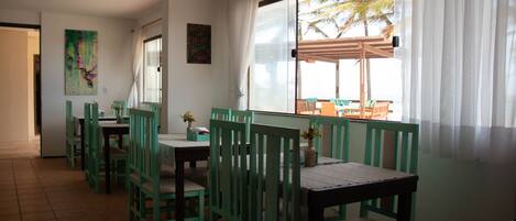 Un comedor con sillas y mesas verdes, un ventanal con vistas a palmeras y un cuadro en la pared.
