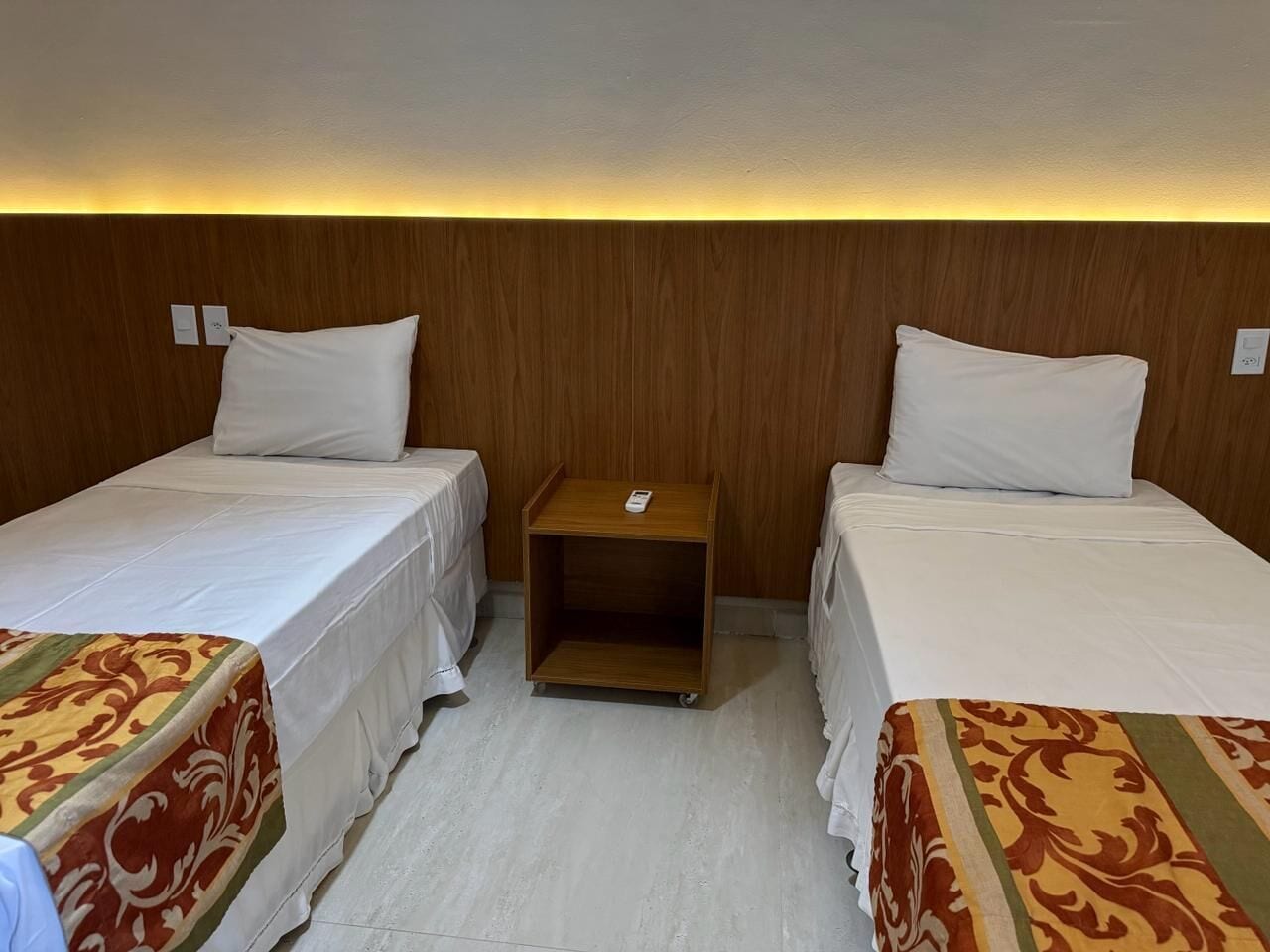 Una camera d'albergo con due letti singoli, un comodino in legno e un pavimento chiaro.