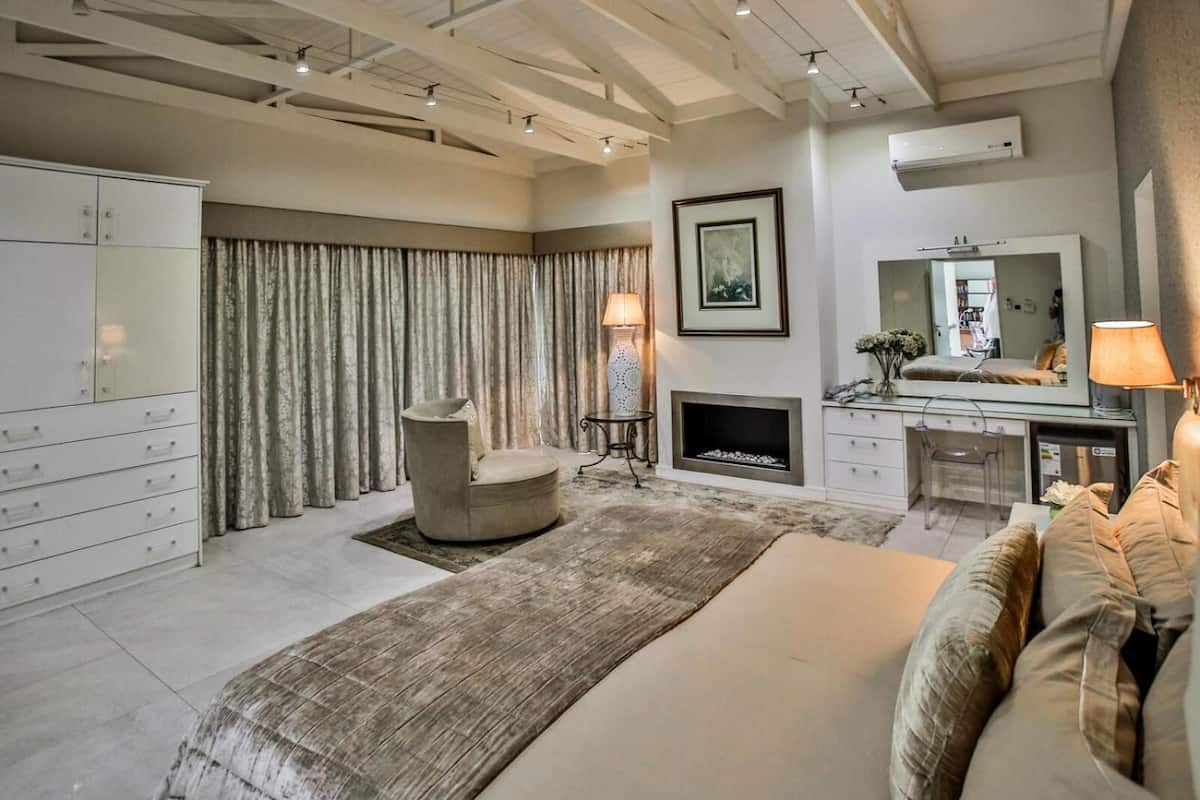 A spacious bedroom with a large bed, a seating area with a chair, a fireplace, and a vanity with a mirror.