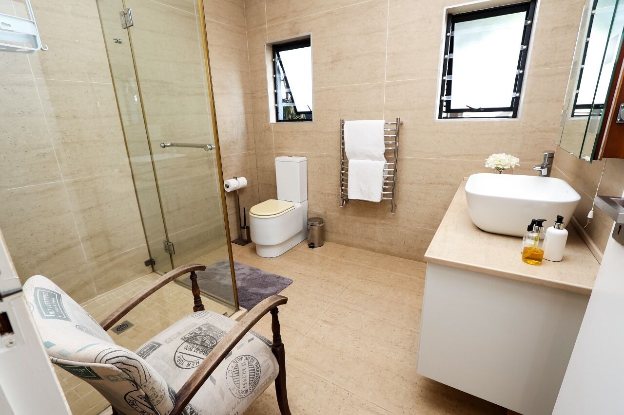 Une salle de bain moderne avec une douche à parois de verre, des toilettes, un lavabo avec un miroir et une chaise dotée d’un coussin à motifs.
