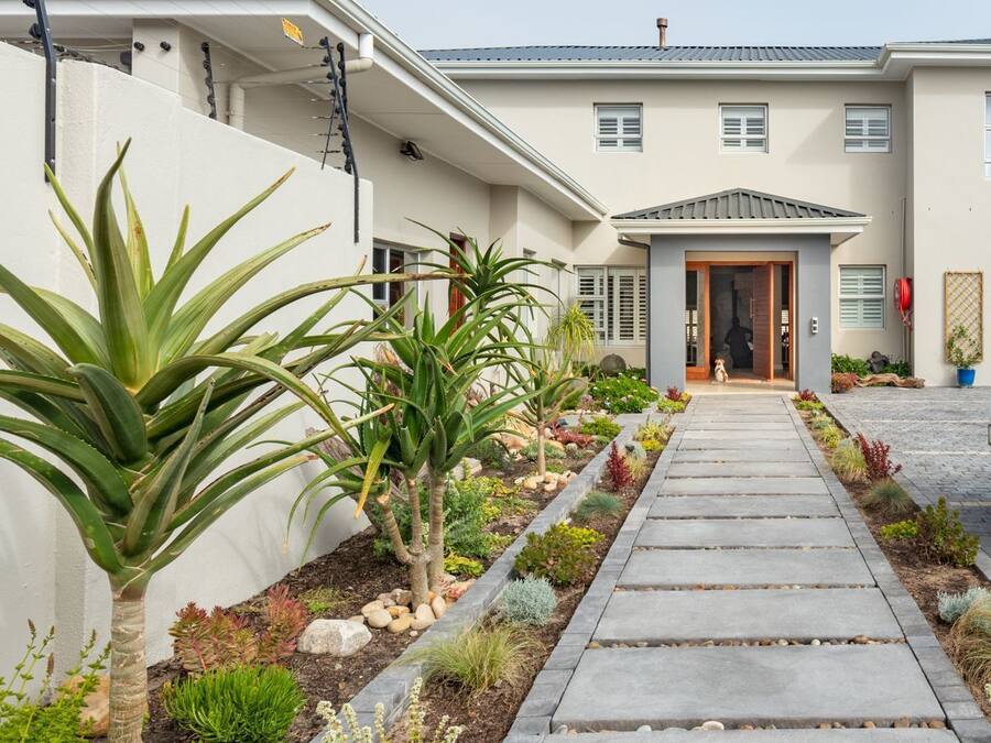 Una casa moderna con entrada pavimentada que conduce a una puerta de madera, rodeada de plantas diversas y un pequeño jardín.