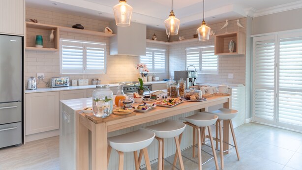 A modern kitchen with white cabinetry, a central island with stools, pendant lighting, and a variety of kitchen appliances and decor. - Bentley's Guesthouse (Cape Town)