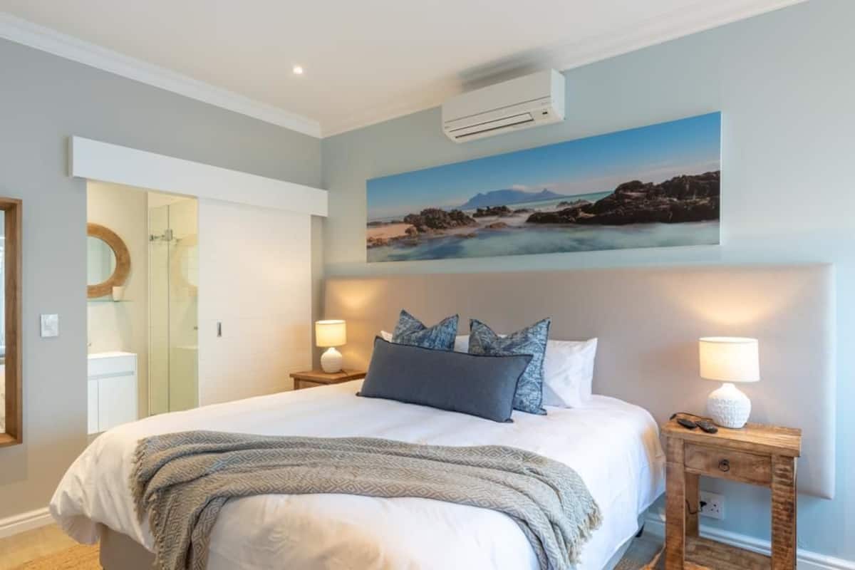 Een slaapkamer met een groot bed, twee nachtkastjes met lampen, een wandairconditioner en een schilderij van een kustlandschap.