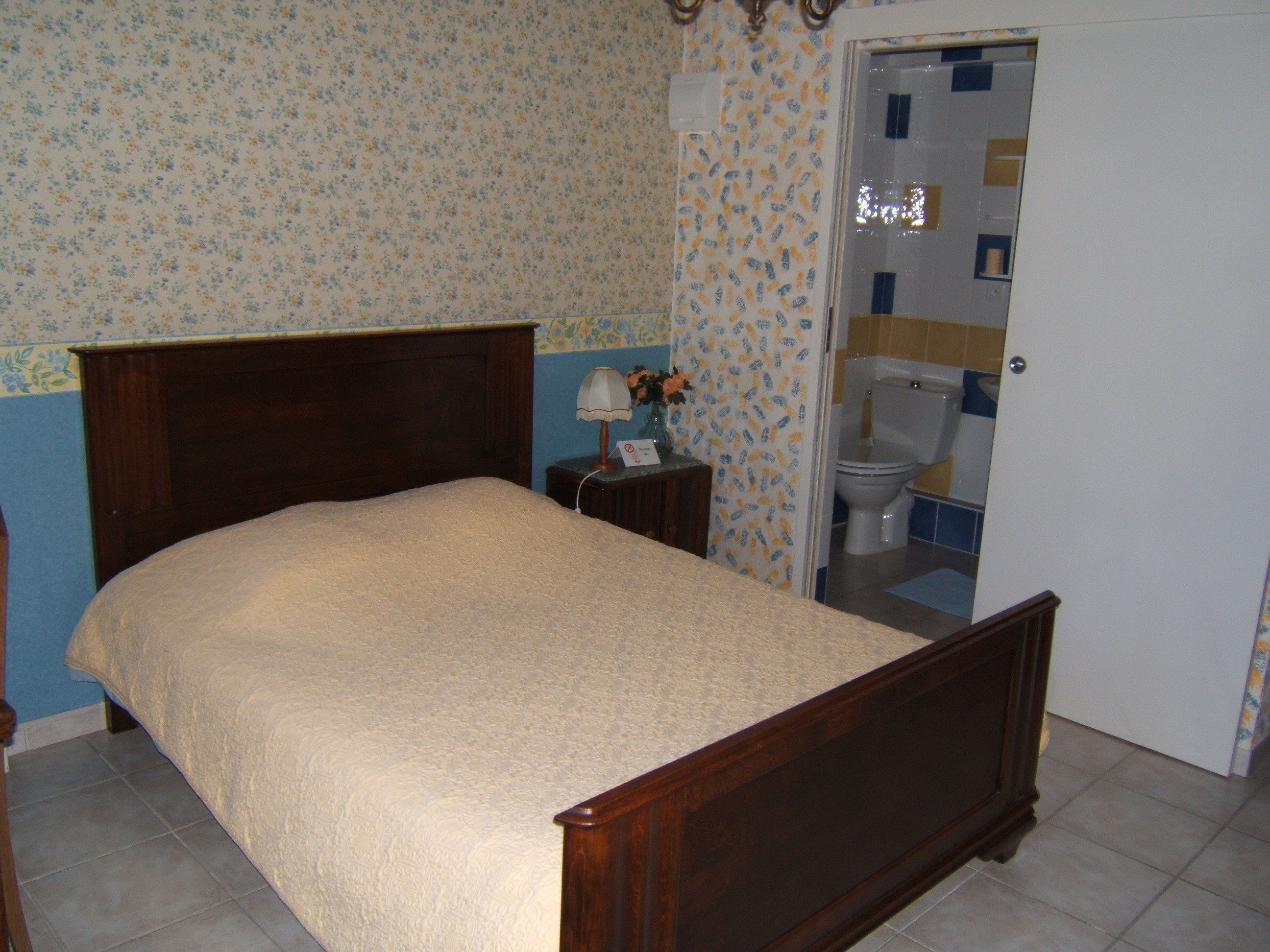 Une chambre à coucher avec un lit en bois, une table de chevet, une lampe et une salle de bain visible à travers une porte ouverte.
