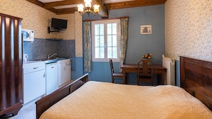 A bedroom with a large bed, a wooden desk, a chair, a television, and a kitchen area with cabinets and a microwave.