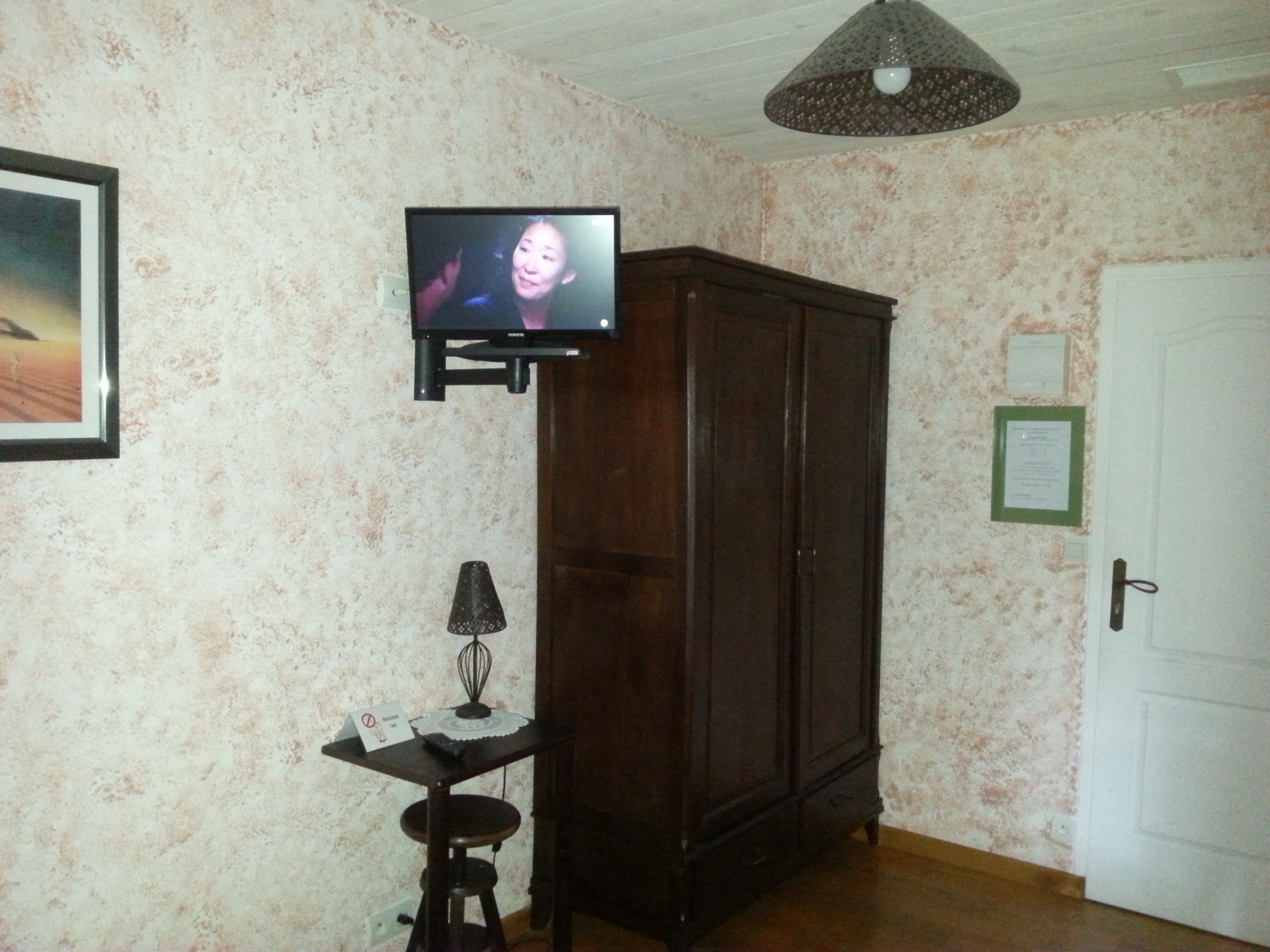 A room with a television mounted on the wall, a wooden cabinet, a small table with a lamp, and a door with a sign.