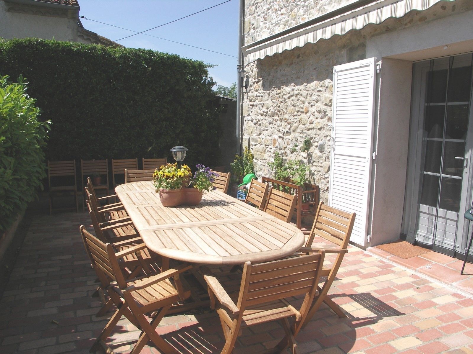 Un patio al aire libre con una mesa y sillas de madera, rodeado de plantas en macetas y un muro de piedra.