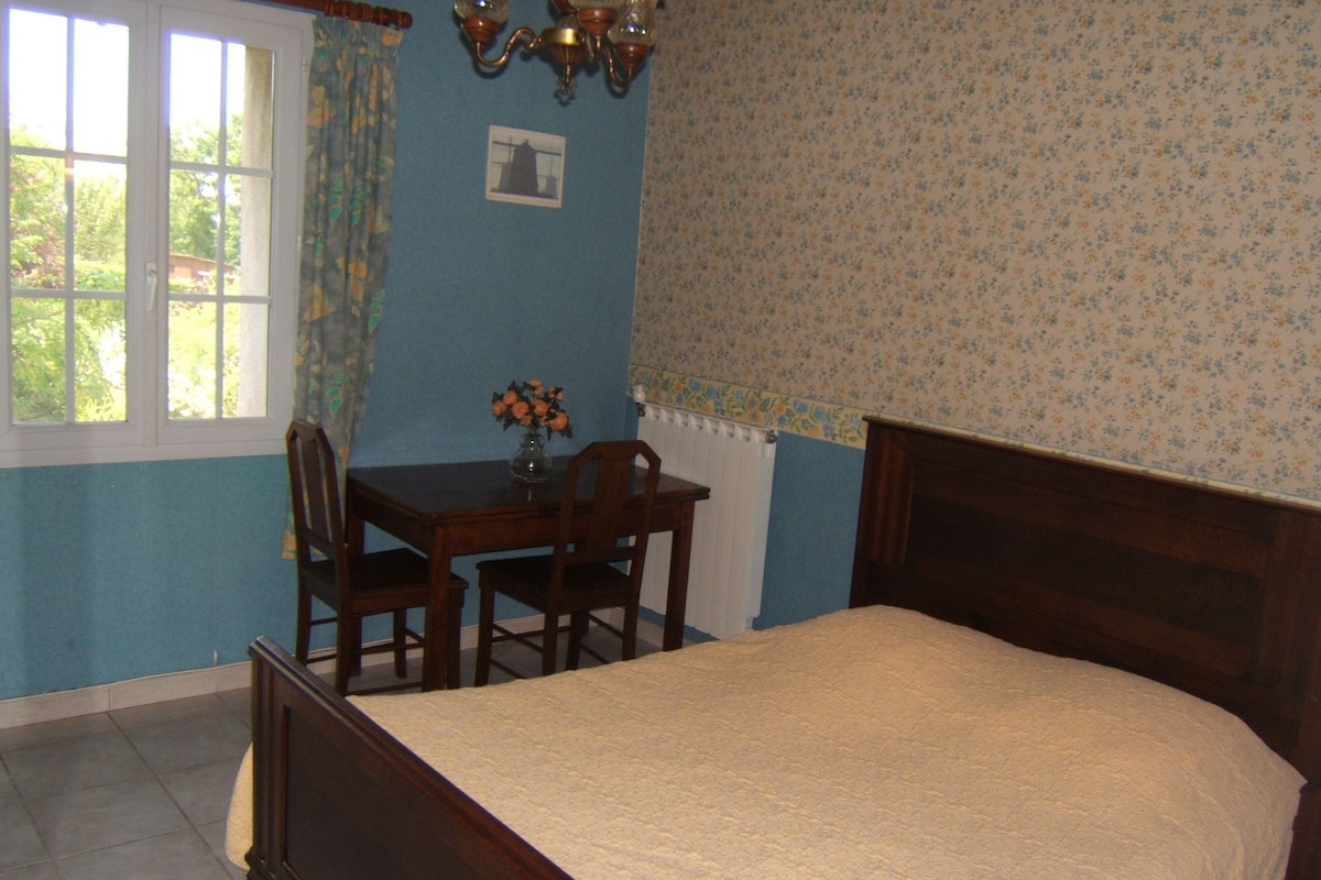 Une chambre avec un lit en bois, une petite table avec des chaises, une fenêtre avec des rideaux et un mur orné d’un motif floral.