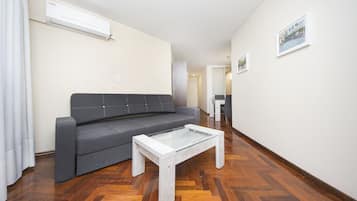 A modern living room with a grey sofa, a glass-top coffee table, wooden parquet flooring, and a white air conditioning unit on the wall.