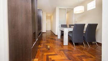 A modern hotel room with a wooden floor, a dining table with chairs, a TV, and a large window.