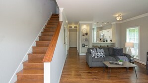 Un vaste espace de vie agrémenté d’un escalier en bois, d’un canapé gris avec des coussins à motifs et d’une cuisine en arrière-plan.