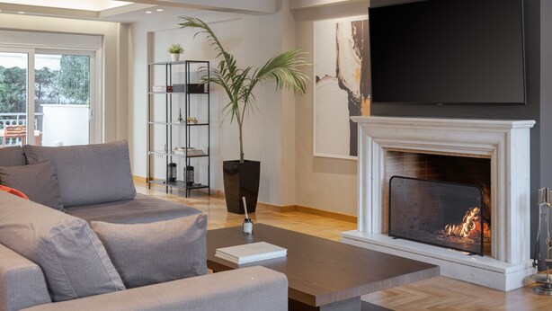 A modern living room with a fireplace, a flat-screen TV mounted above it, and a large window providing a view of the outdoors. - Athenian Grand Riviera Villa (Vari-Voula-Vouliagmeni)