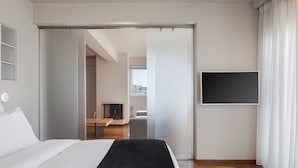 A modern hotel room with a bed, a flat-screen TV, and a view of the city through the window. - Athenian Grand Riviera Villa (Vari-Voula-Vouliagmeni)