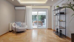 A modern living room with a white sofa, wooden flooring, a glass door leading to an outdoor balcony, and a black metal shelf unit. - Athenian Grand Riviera Villa (Vari-Voula-Vouliagmeni)