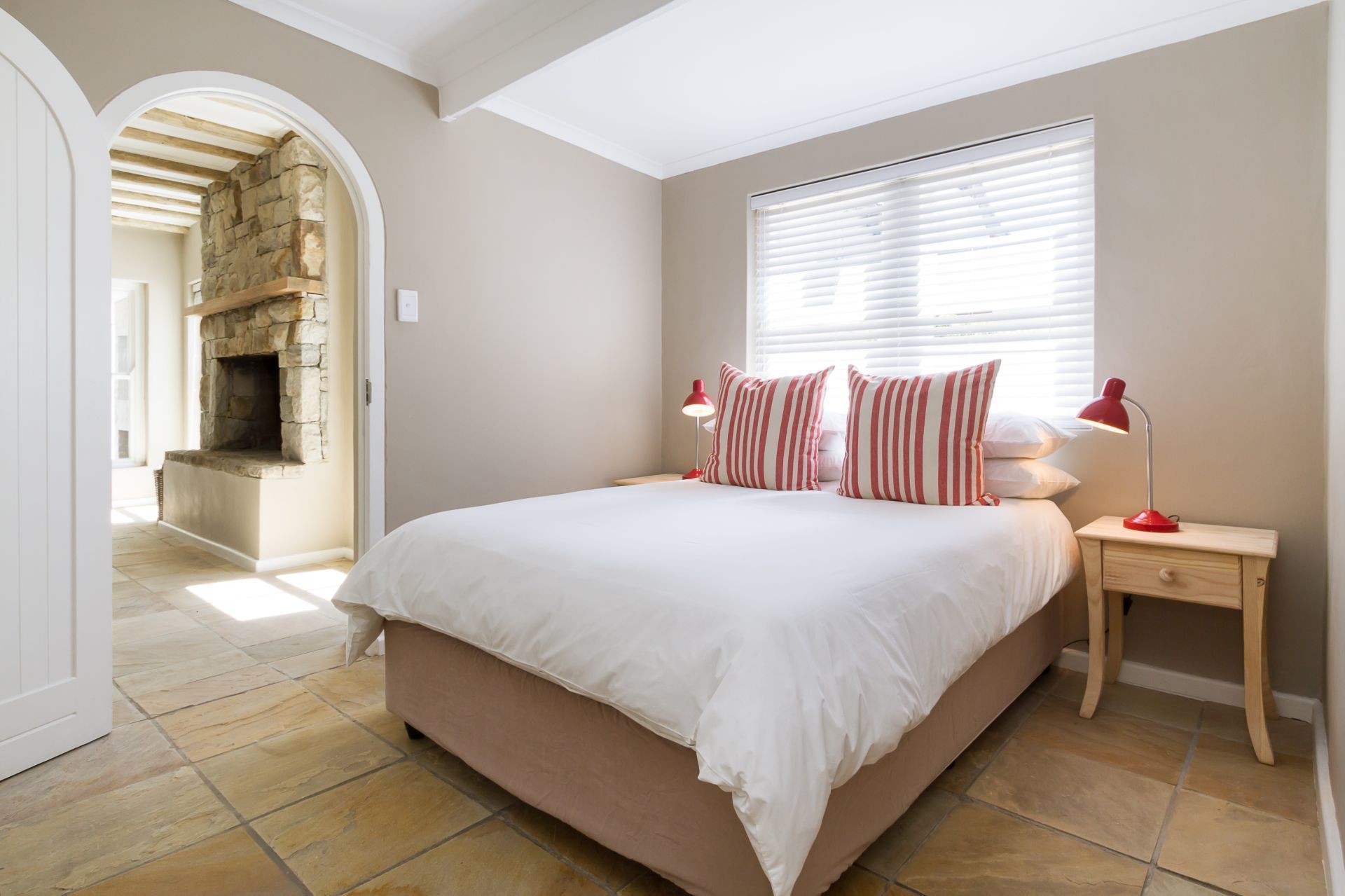Quarto com lareira de pedra, cama grande com roupa de cama branca, duas luminárias vermelhas e janela com persianas.