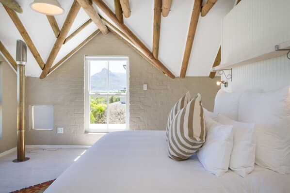 A bedroom with a large bed, wooden beams, a view of greenery and mountains, and a window.