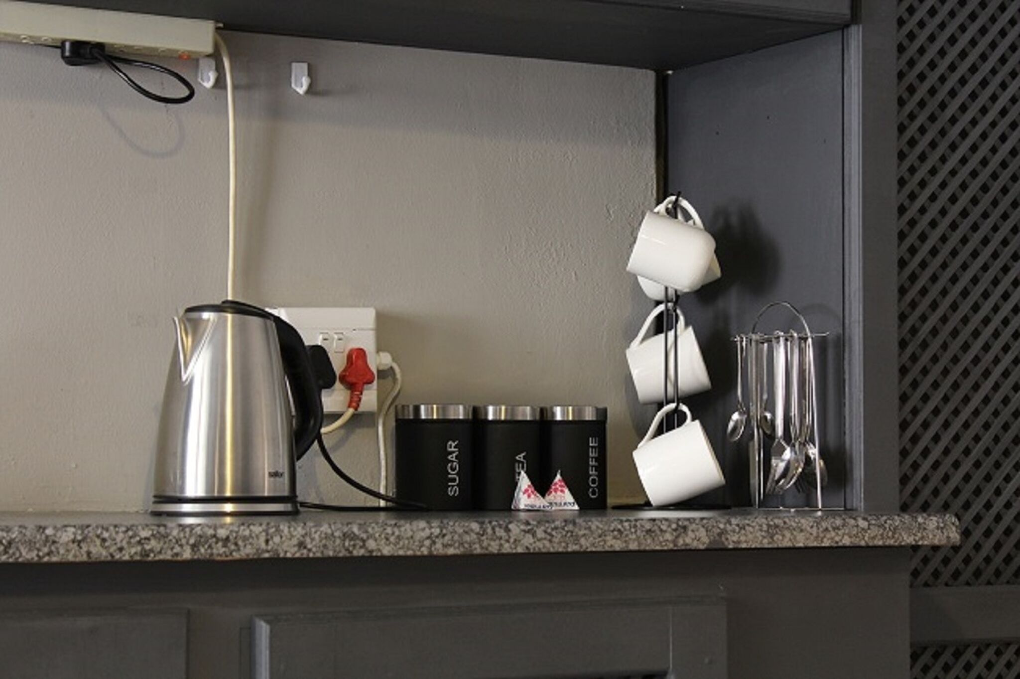 A kitchen counter with a kettle, sugar, tea, and coffee containers, along with a set of white mugs and a glass.