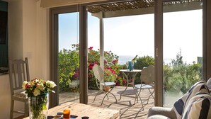 A room with a wooden table, a vase of flowers, and a view of the sea through large glass doors. - Watercolours Studios (Cape Town)