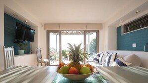 A modern living room with a dining table in the center, a large window, and a television mounted on the wall. - Watercolours Studios (Cape Town)