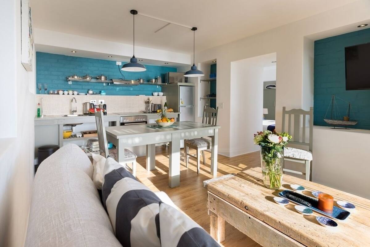 A modern kitchen with a blue backsplash, white cabinets, and stainless steel appliances. There is a dining table with chairs and a vase of flowers on the table.