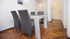 A dining area with a glass table and four grey chairs, a wooden herringbone floor, and a door in the background. - Obispo Oro Flat ZX1 (Córdoba)