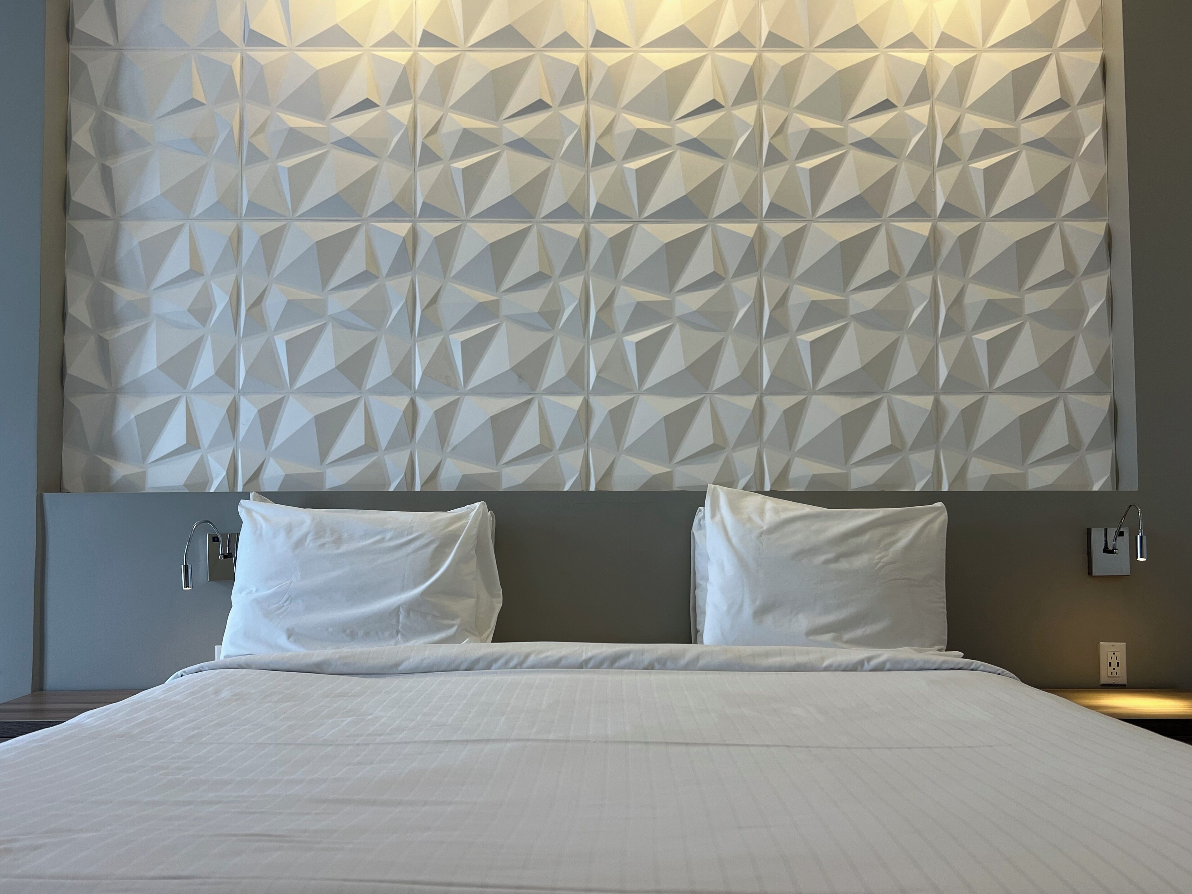 Una habitación de hotel moderna con un cabecero de estampado geométrico, una cama bien tendida con sábanas blancas y una mesita de noche con lámpara.