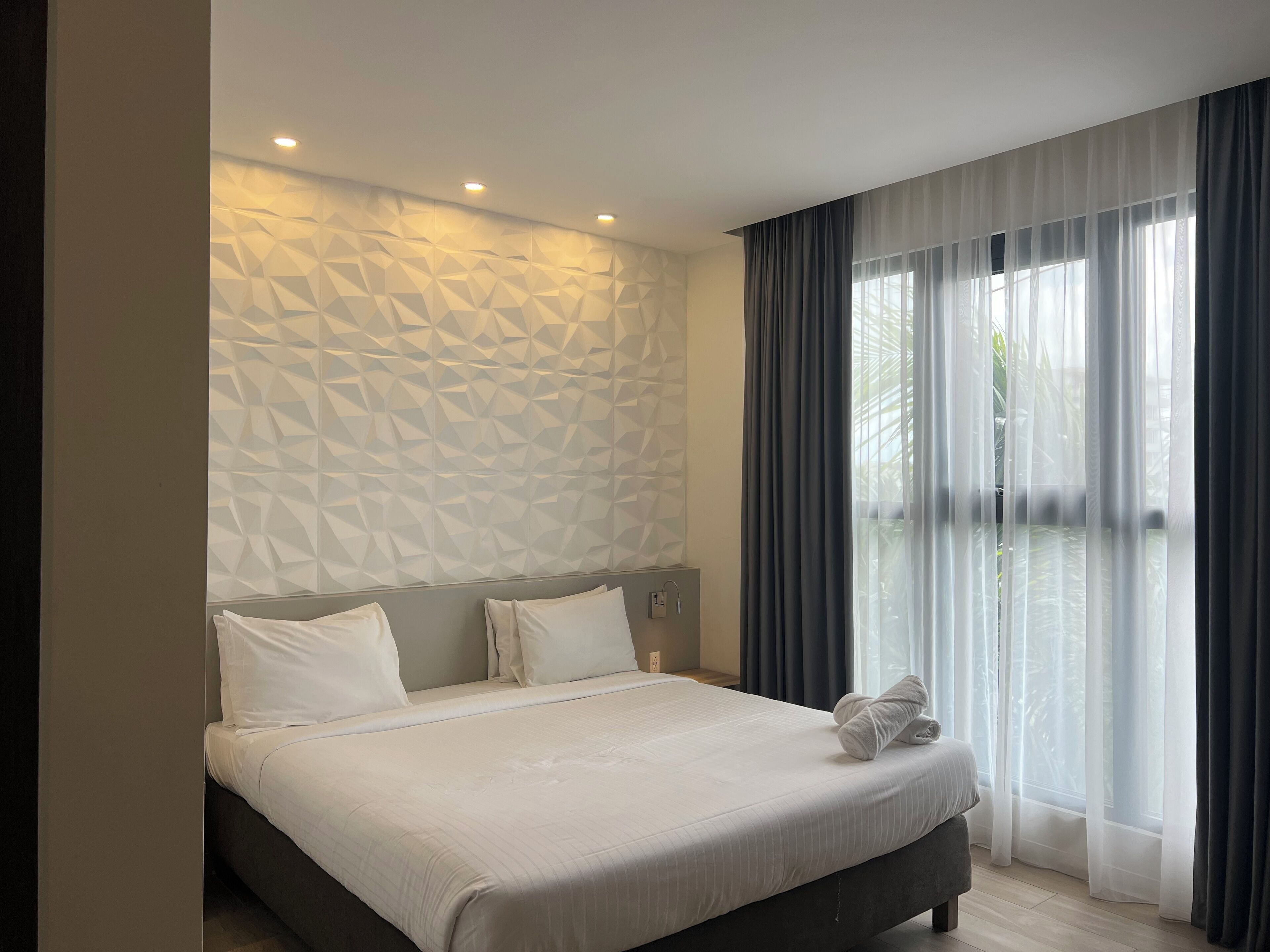 Une chambre d’hôtel moderne avec un mur orné d’un motif géométrique, un grand lit avec du linge de maison blanc et une vue sur la verdure à travers des rideaux transparents.