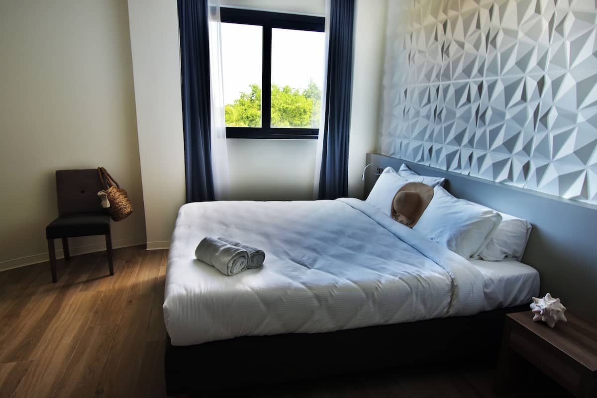 Un lit bien fait, avec du linge de lit blanc, une table de chevet en bois, une chaise avec un sac en osier, et un mur orné d’un motif géométrique.
