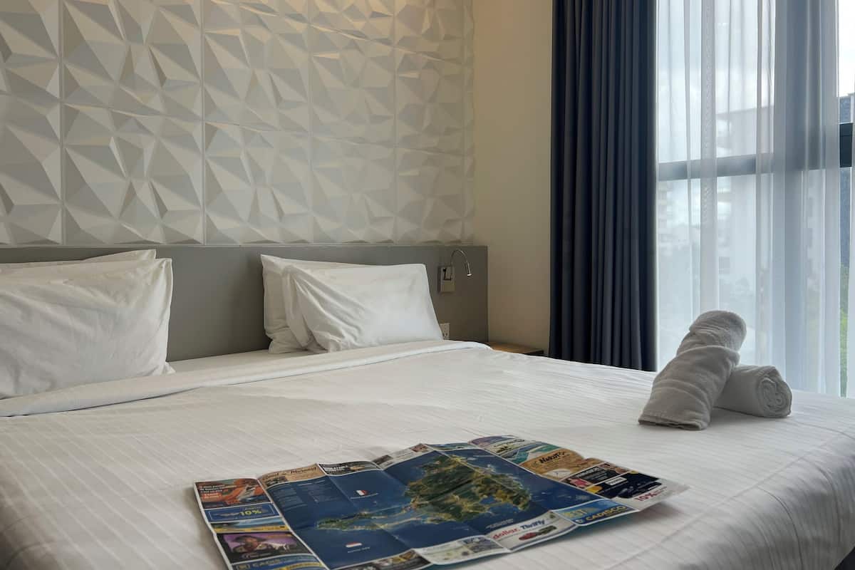 Une chambre d’hôtel avec un mur orné d’un motif géométrique, un lit soigneusement fait avec une serviette pliée, et une fenêtre avec des rideaux légers.