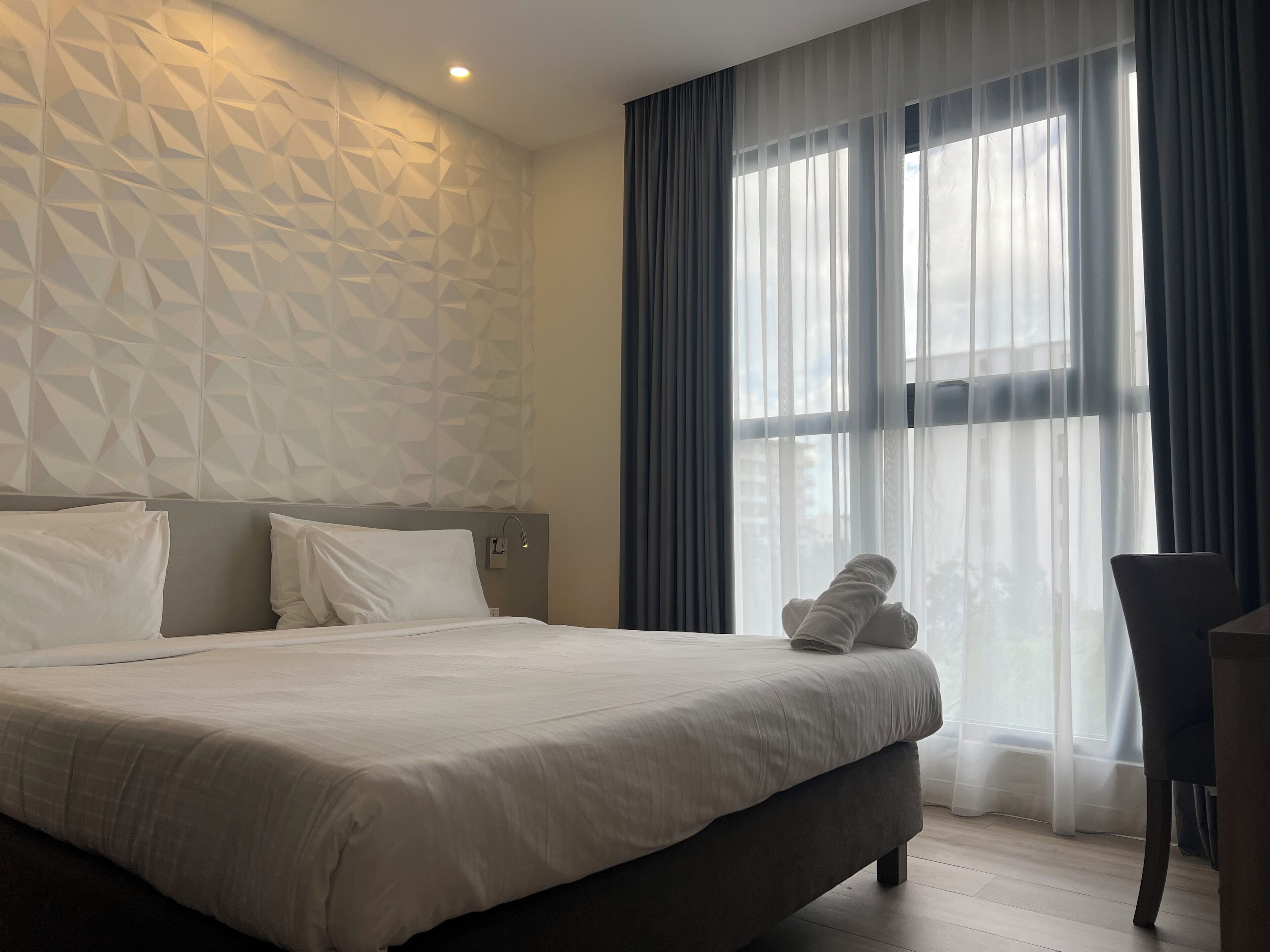 Ein modernes Hotelzimmer mit Wand im geometrischen Muster, einem großen Bett mit weißen Leinen und Blick ins Freie durch transparente Vorhänge.