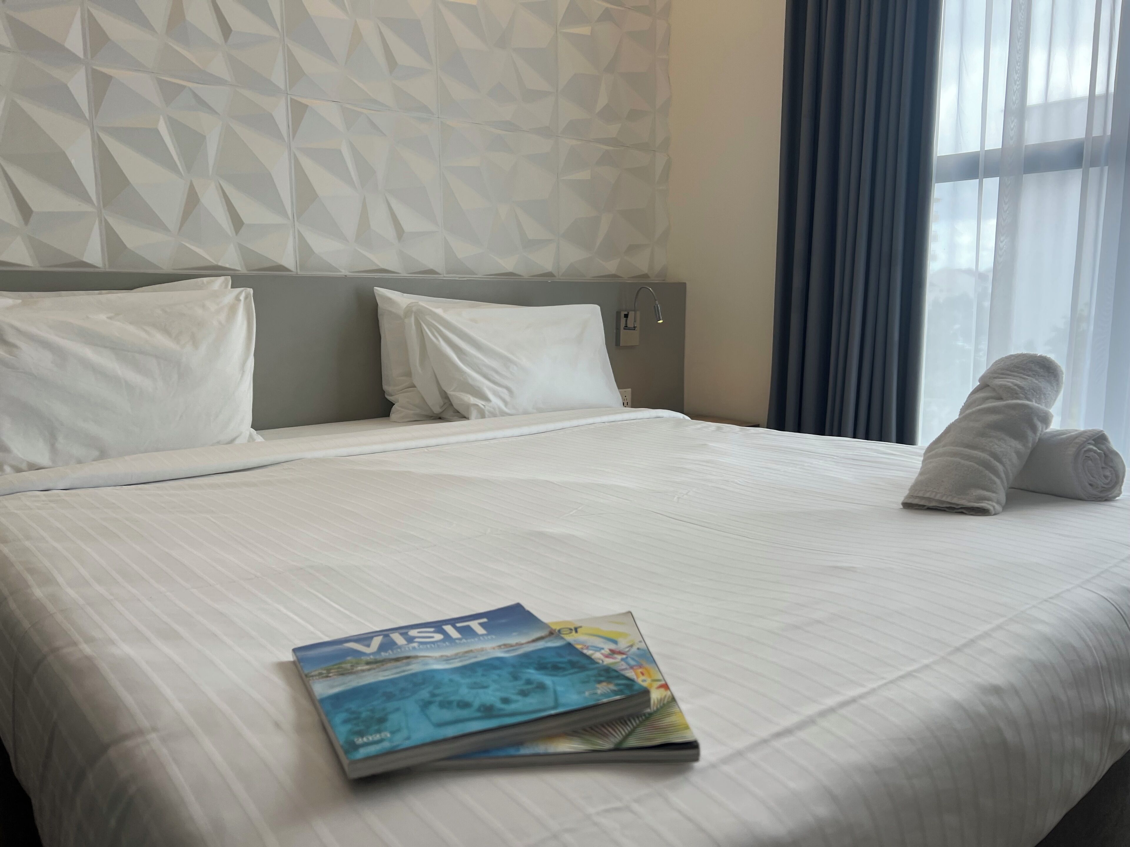 Habitación de hotel con una cama bien hecha, una almohada, una toalla enrollada y un folleto de 'Visit' sobre la cama.