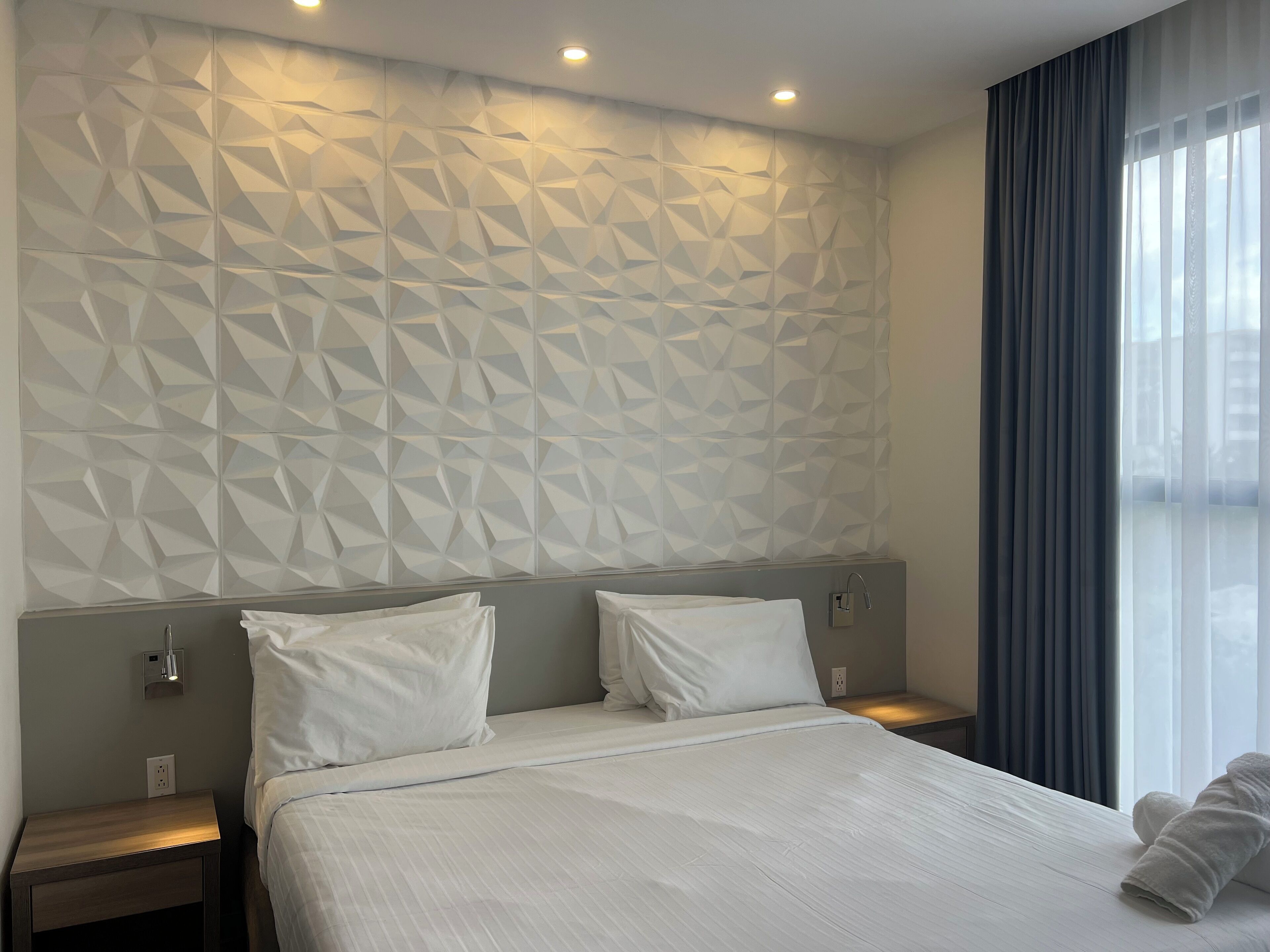 Habitación de hotel con cabecera de diseño geométrico, cama grande con ropa blanca, dos mesitas de noche con lámparas y vistas al exterior a través de cortinas traslúcidas.