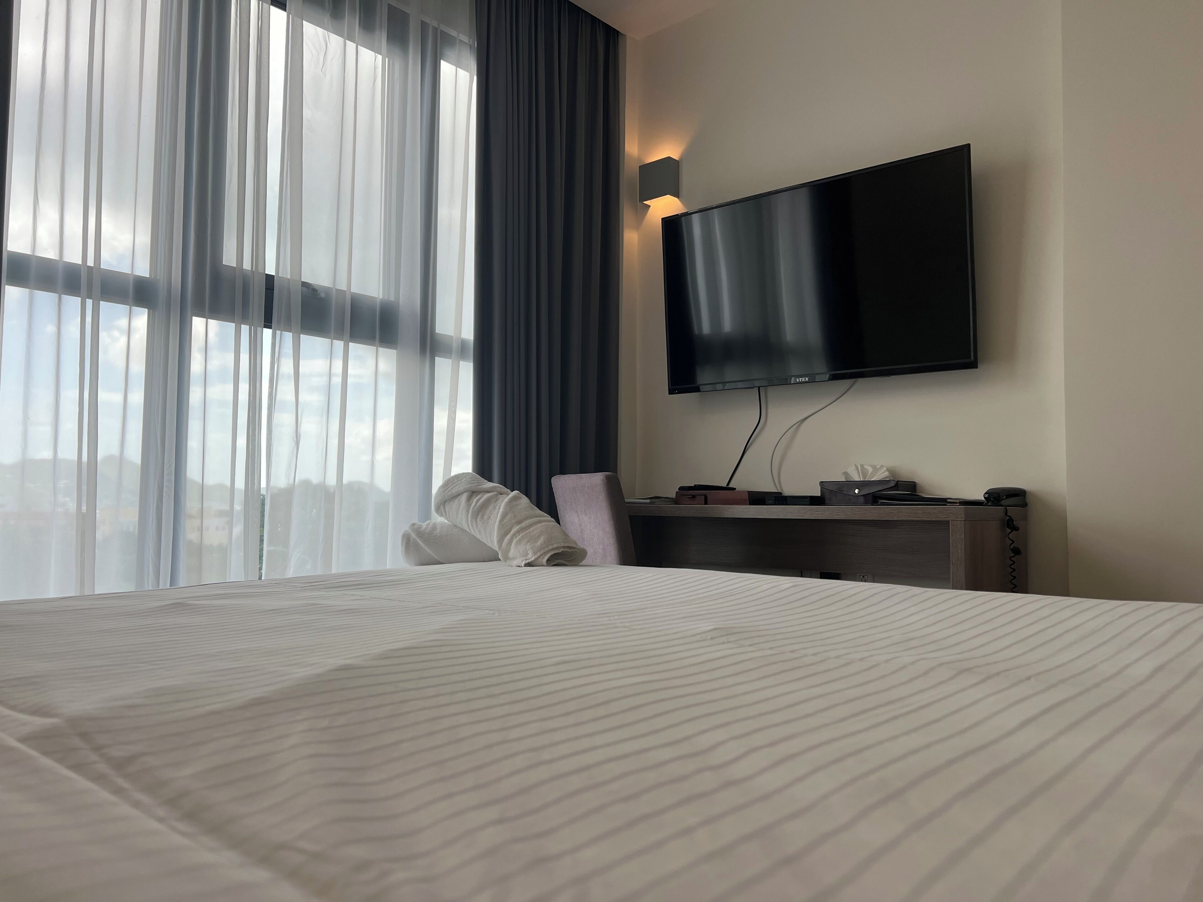 A hotel room with a large bed, a flat-screen TV mounted on the wall, a desk with a chair, and a window with sheer curtains.