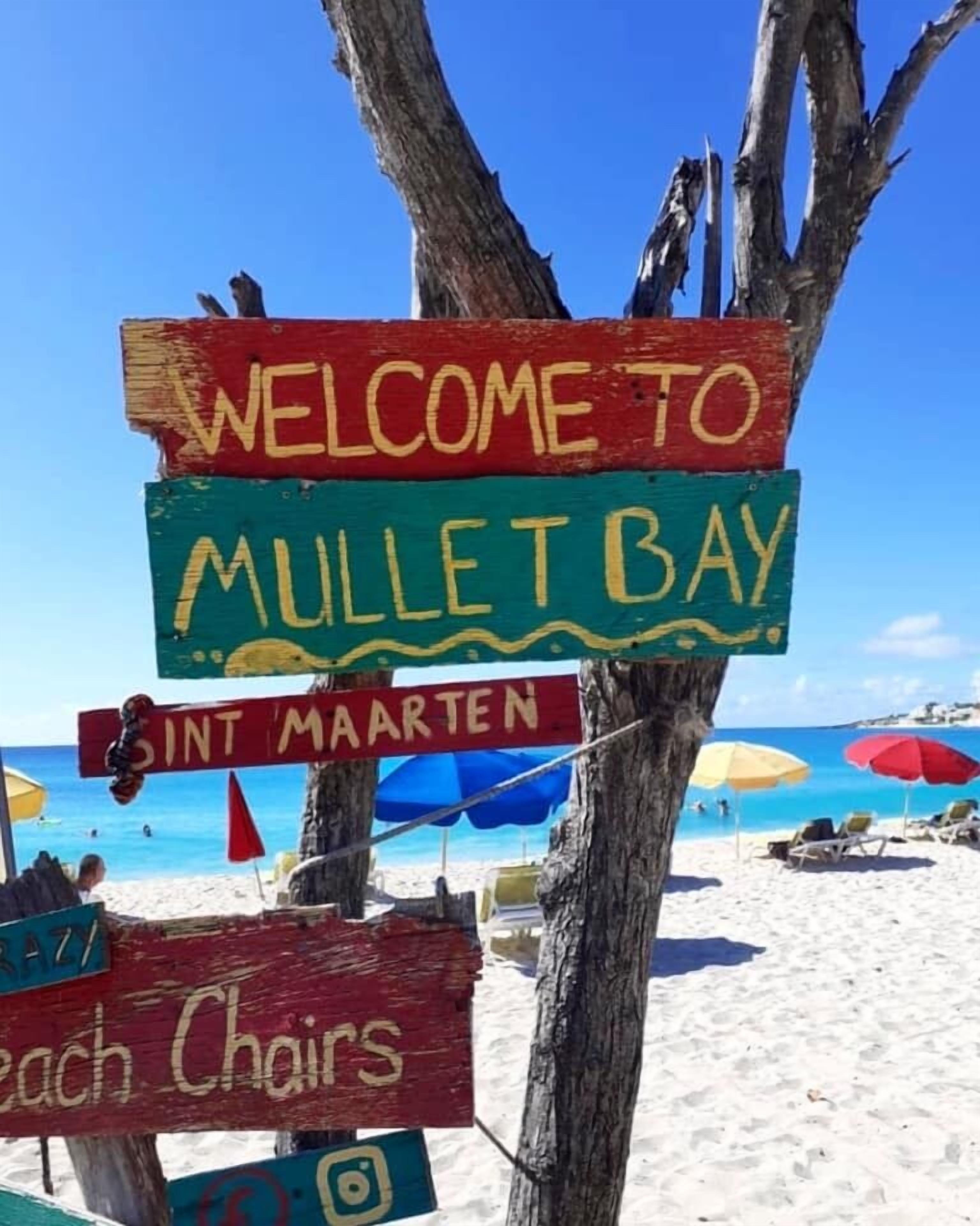 Un panneau sur la plage sur lequel on peut lire « BIENVENUE À MULLET BAY » et « SINT MAARTEN », avec des parasols et des chaises de plage en arrière-plan.