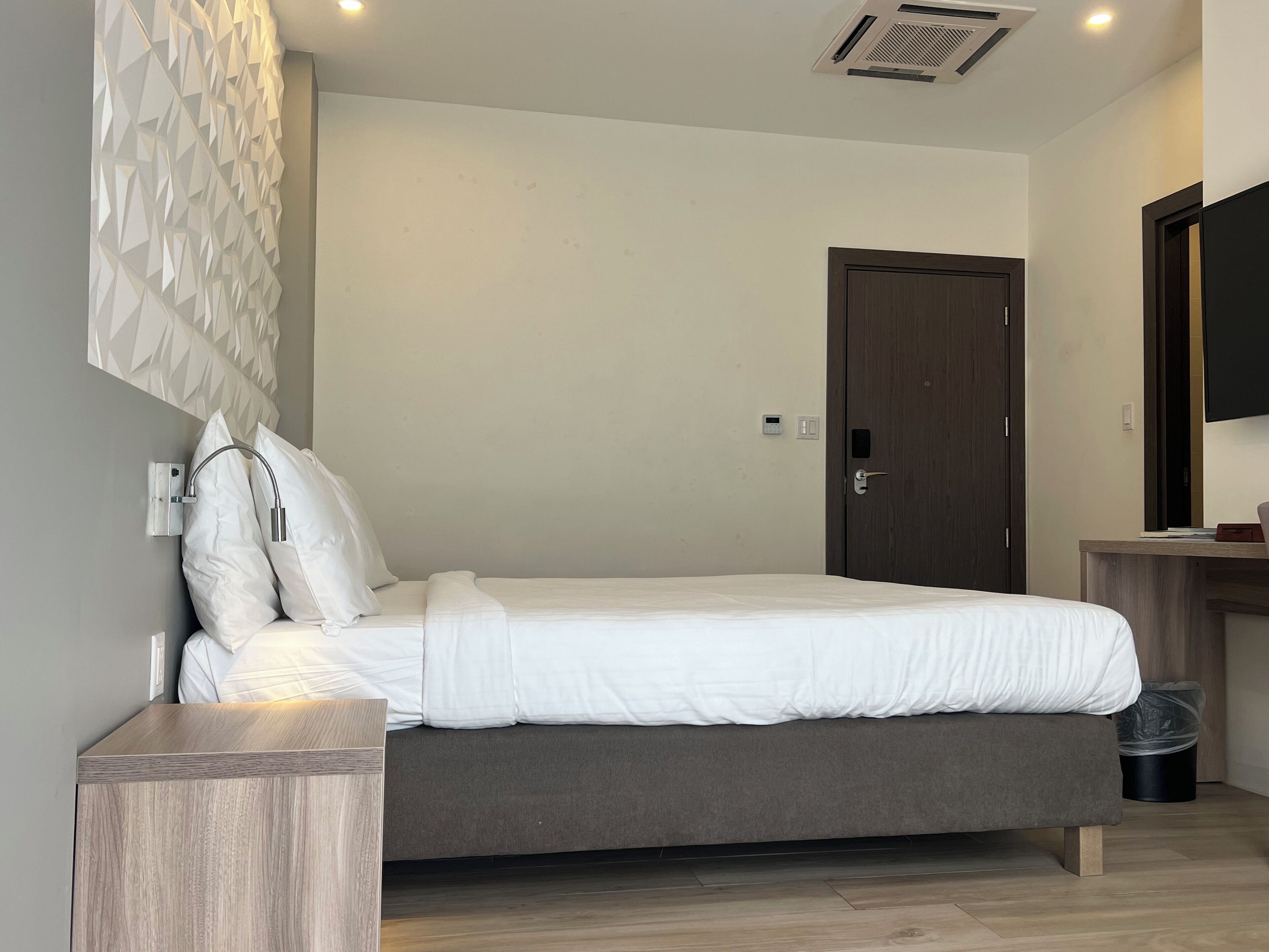 Un dormitorio moderno con una cama, una mesita de noche de madera, una lámpara fijada a la pared, una puerta y un televisor en la pared.