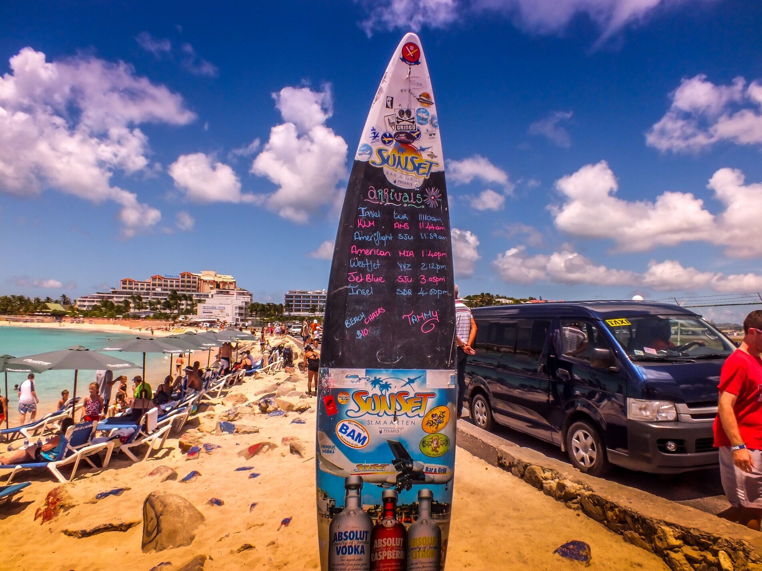 A beachside bar with a chalkboard menu listing drinks and prices, a surfboard, and a view of the beach and ocean.