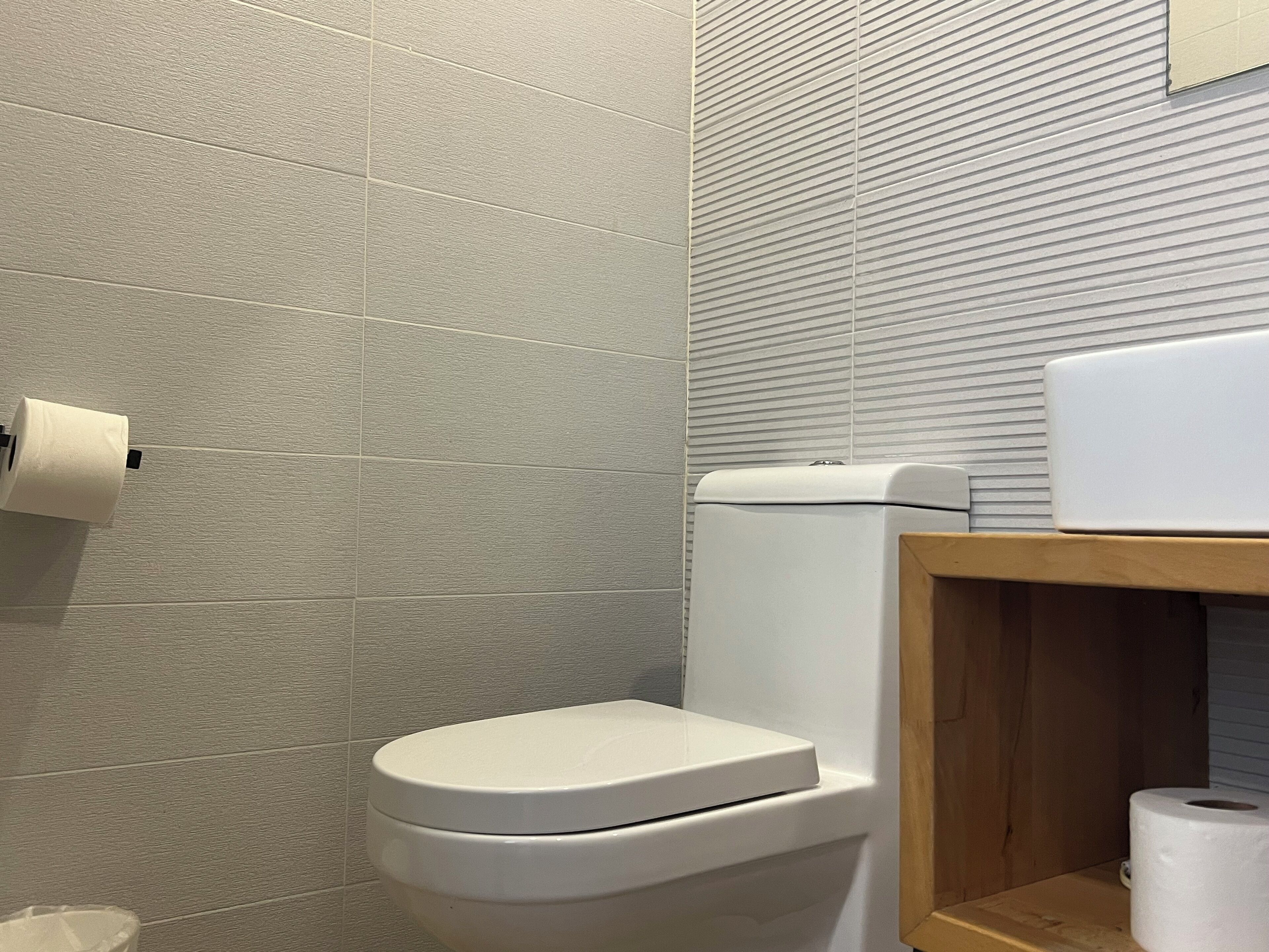 A bathroom with a toilet, a toilet paper holder, and a wooden shelf with a roll of toilet paper.