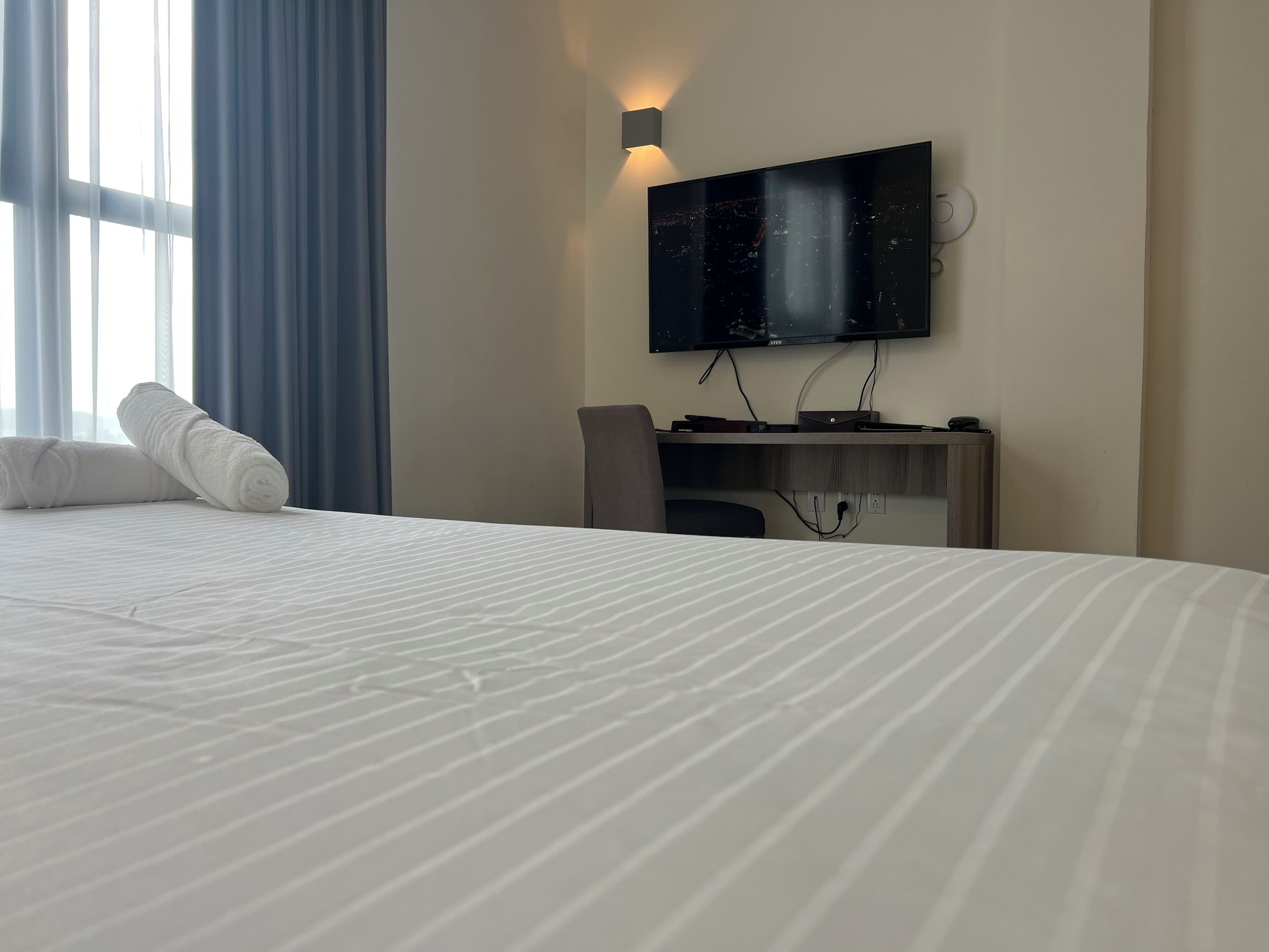 Habitación de hotel con una cama grande, un escritorio con computadora, un televisor de pantalla plana montado en la pared y una ventana con cortinas azules.